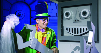 hombre vestido con chaqueta verde y morada, chistera, gafas al lado de un robot gigante